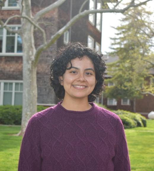 Photo of Abi: short curly dark hair, smiling, wearing a deep purple knit sweater. Background includes green grass, a thin tree with few leaves, and a brick building.