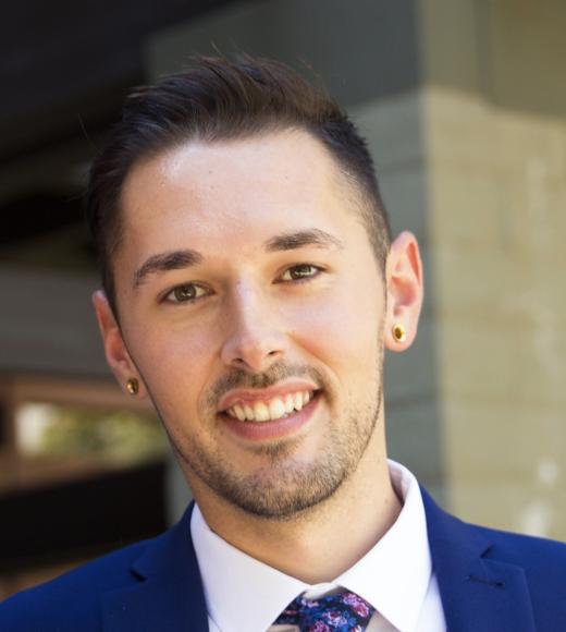 Smiling young man in blue suit and floral tie, photographed against a neutral background.