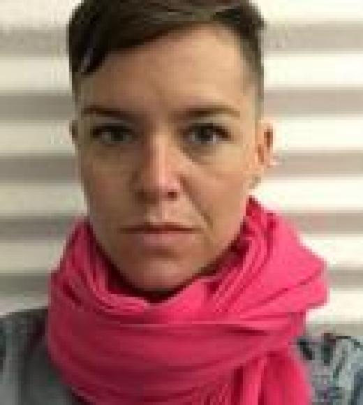 Selfie of Megan Bayles wearing a pink scarf and looking into the camera. She has short hair and is standing in front of a striped background. 