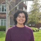Photo of Abi: short curly dark hair, smiling, wearing a deep purple knit sweater. Background includes green grass, a thin tree with few leaves, and a brick building.