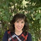 Photo of GSW Chair Kathryn Olmsted - she is smiling and sitting in front of a leafy green background. She is wearing a navy blue sweater with a red and white pattern. 
