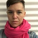 Selfie of Megan Bayles wearing a pink scarf and looking into the camera. She has short hair and is standing in front of a striped background. 